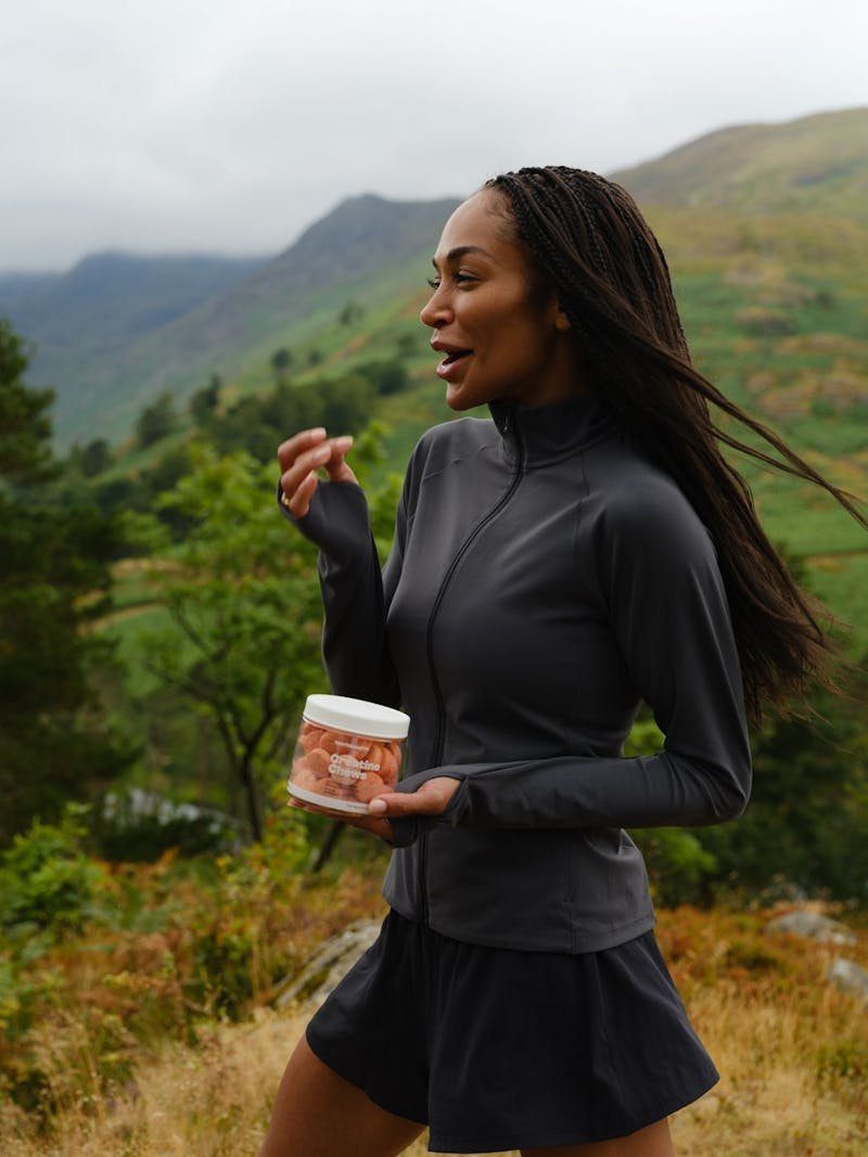 A woman in grey activewear walking through green hills, holding a tub of Creatine Chews