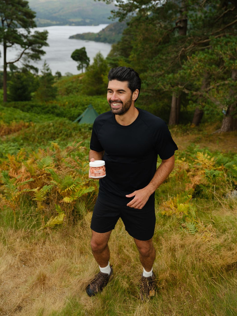 A man smiling on a hillside walk, holding a tub of Creatine Chews